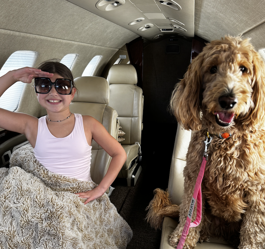 Izzy, a young girl with sunglasses saluting, and her dog sitting inside a private jet, smiling.